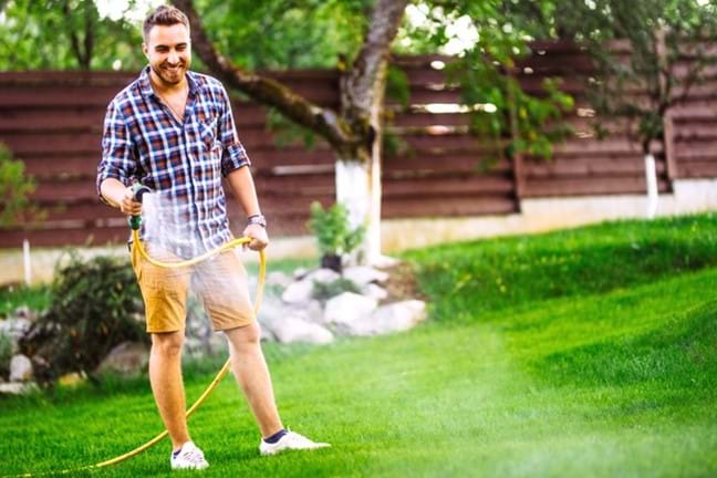 Man wearing flannelette shirt and shorts watering a lush with nice garden in background