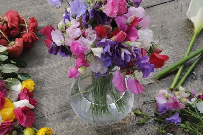 a glass vase full of fresh-cut pink, purple and red sweet pea flowers sitting on a timber table where there is various other cit flowers laying on bench